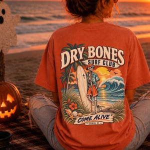 Peut inclure: Un t-shirt couleur rouille avec un motif au dos représentant un squelette tenant une planche de surf sur une plage. Le texte indique DRY BONES SURF CLUB et COME ALIVE EZEKIEL 37. L'illustration inclut des palmiers, un coucher de soleil et des vagues.