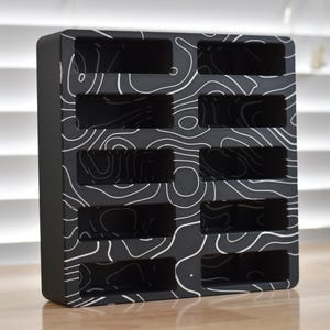 May include: A black organizer with a white topographic pattern. The organizer has multiple rectangular slots. The product is made of a solid material and is designed to hold small items. The design is modern and minimalist.