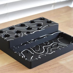 May include: A black organizer with a white topographic pattern. The organizer has two levels with multiple circular holes and a rectangular tray. The design is modern and geometric.