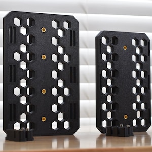 May include: Two black, rectangular panels with a honeycomb pattern and gold-colored fasteners. The panels have a series of hexagonal cutouts and vertical slots. Small black bases are at the bottom of each panel. The panels are on a wooden surface.