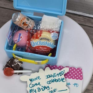 Peut inclure: Une petite boîte à lunch bleue avec une poignée jaune, contenant des collations et une boisson. Comprend une canette d'OLIPOP, une collation rose Hostess, un sandwich Smucker's Uncrustables et une serviette. Des cartons d'invitation d'anniversaire sont également présents.