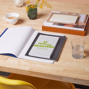 Puede incluir: Un cuaderno abierto con encuadernación azul junto a una pila de libros y un vaso de agua sobre una mesa de madera. El libro superior tiene el texto "LE DROIT DES OBLIGATIONS" en verde. Una taza de té y un jarrón con flores amarillas también están sobre la mesa.