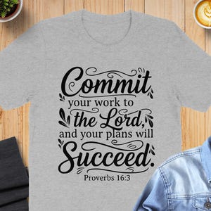 May include: A light gray t-shirt with the text "Commit your work to the Lord, and your plans will Succeed. Proverbs 16:3" in black script. The shirt is displayed on a wooden surface.