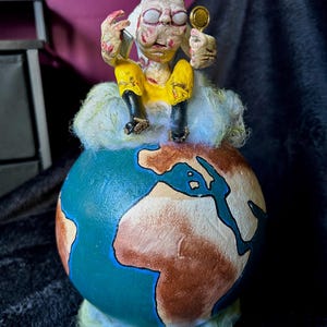 May include: A quirky figurine featuring a seated character on a globe. The figure, with a pale complexion and yellow attire, holds a small, circular item. The globe is painted with continents and rests on a cloud-like base, making it a unique decorative item.