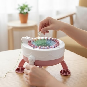 May include: A white and pink knitting machine with a circular design. The machine has a pink base and legs, with a white top and a circular knitting area. The machine is being used with colourful yarn.