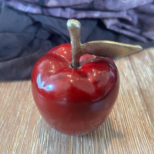 May include: A glossy, red apple sculpture with a metallic stem and leaf. The apple has a smooth, reflective surface. The stem is silver-colored, and the leaf is gold-toned. This decorative piece is a fruit-themed home decor item.