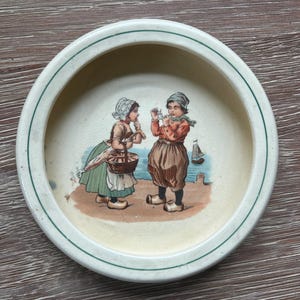 May include: A vintage ceramic bowl with a cream-colored interior and a green-lined rim. The bowl's center features a colorful illustration of two children in traditional Dutch attire, standing on a beach with a sailboat in the background.