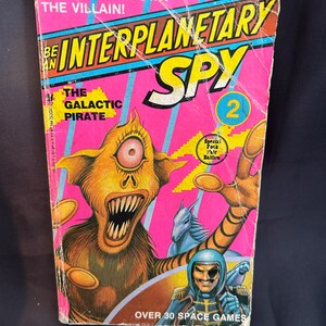 May include: A vintage "Be an Interplanetary Spy" book cover. The cover has a pink background with the title in yellow and blue. An orange alien with a large eye and teeth is featured, along with a space pilot and the text "Over 30 Space Games."