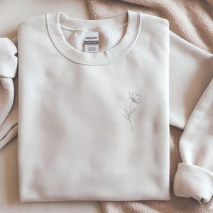 Peut inclure: Sweat-shirt crème avec un motif floral minimaliste noir sur le devant. Le dessin d'une fleur est simple. Le sweat-shirt a un col rond et des manches longues, idéal pour un look décontracté.