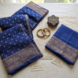 May include: A display of blue and gold Banarasi saree fabric, featuring detailed patterns. The image includes two gold bangles, a wooden block with a floral design, and two small cards with text.