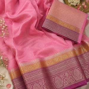 Könnte beinhalten: Ein pinkfarbener Sari mit aufwendigen gold- und magentafarbenen Bordüren. Der Stoff drapiert sich elegant und zeigt ein passendes Blusenteil mit ähnlichen Details. Das Design des Saris weist florale und geometrische Muster auf.