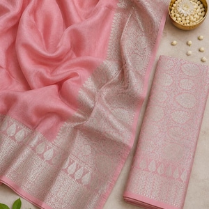 May include: A draped, light pink fabric with a silver patterned border and a matching folded fabric piece. A small gold bowl with white flowers sits near the fabric. The fabric has a soft, flowing texture.