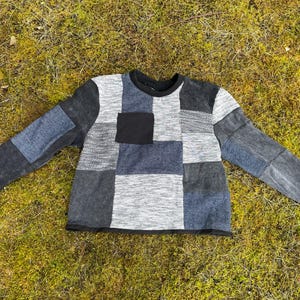 Könnte beinhalten: Ein Patchwork-Langarmshirt in Grau-, Schwarz- und Blautönen. Das Shirt hat einen Rundhalsausschnitt, eine schwarze Tasche und schwarze Manschetten. Das Design besteht aus verschiedenen zusammengenähten Stoffquadraten.