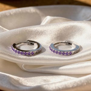 May include: A pair of silver hoop earrings with a row of small, purple gemstones. The earrings are displayed on a white, silky fabric, catching the light and showcasing the jewelry's sparkle. The design is simple yet elegant.