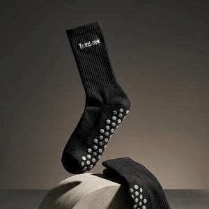 Pode incluir: Um par de meias pretas de cano médio com textura canelada e o texto branco Thirdlove bordado no punho superior. As solas das meias são cobertas por um padrão de pequenos pontos de aderência circulares de silicone cinza claro.