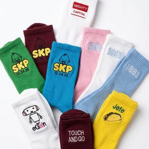 Puede incluir: Una colecci&oacute;n de nueve calcetines de ca&ntilde;a media en varios colores, incluyendo blanco, verde, granate, azul, rosa y amarillo. Cada calcet&iacute;n presenta dise&ntilde;os bordados &uacute;nicos y textos como MIGHTY CAPITAL, SKP 12.24.26, 1881, eGain, TOUCH AND GO y Jefe con un taco.