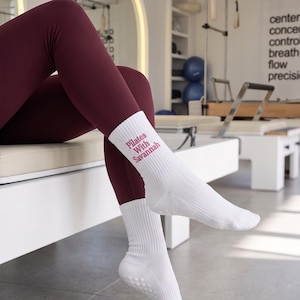 Op de afbeelding: Een paar witte enkelsokken met de tekst "Pilates With Savannah" in roze letters aan de zijkant. De sokken hebben een geribbelde textuur en kleine witte antislipnoppen op de zolen, gedragen met een donkerbordeauxrode legging.
