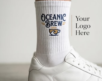 Puede incluir: Un calcetín blanco de canalé con un diseño bordado que incluye el texto azul oscuro Oceanic Brew sobre un icono de taza de café dorada y azul con un gráfico de olas. El calcetín se lleva con una zapatilla blanca de cuero sobre una superficie.