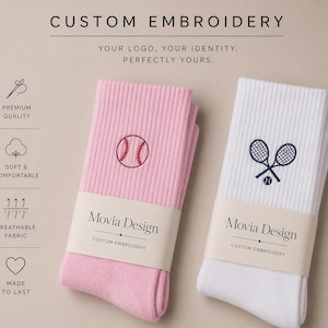 Könnte beinhalten: Zwei Paar individuell bestickte Socken. Eine rosa Socke mit Baseball-Design und die andere weiße Socke mit Tennisschläger und Ball. Die Socken tragen den Text "Movia Design" und "Custom Embroidery" auf dem Etikett.
