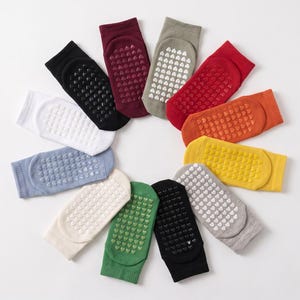Puede incluir: Una disposición circular de once calcetines tobilleros en varios colores sólidos, incluyendo negro, blanco, rojo, naranja, amarillo, verde, azul y gris. Cada calcetín presenta un patrón de agarre texturizado en la suela con pequeñas formas de corazón.