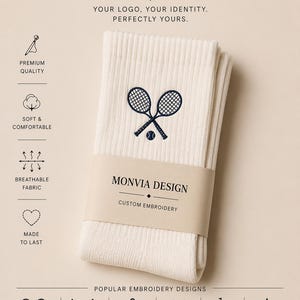 Puede incluir: Calcetines acanalados de color blanco roto con un bordado azul oscuro de dos raquetas de tenis y una pelota. Una banda de papel con la etiqueta MONVIA DESIGN CUSTOM EMBROIDERY envuelve el centro. La imagen incluye iconos de caracter&iacute;sticas.