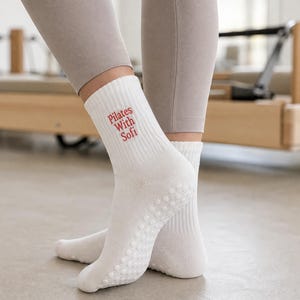 以下が含まれることがあります： リブ編みのカフと側面に赤い文字で「Pilates With Sofi」とプリントされた白いクルー丈のソックス。ソックスの足裏には、運動中のトラクションを高めるための小さな白い円形のシリコン製グリップドットが施されています。