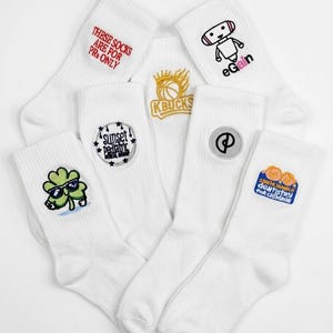 Puede incluir: Un conjunto de seis calcetines blancos con diversos dise&ntilde;os bordados, que incluyen un robot con el texto eGain, un tr&eacute;bol con gafas de sol, un logotipo de baloncesto y textos como These Socks Are For PRs Only y Santa Monica Dentistry.