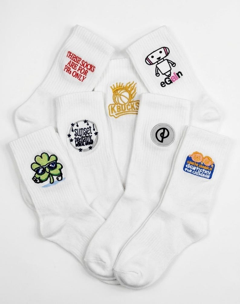 Puede incluir: Un conjunto de seis calcetines blancos con diversos dise&ntilde;os bordados, que incluyen un robot con el texto eGain, un tr&eacute;bol con gafas de sol, un logotipo de baloncesto y textos como These Socks Are For PRs Only y Santa Monica Dentistry.