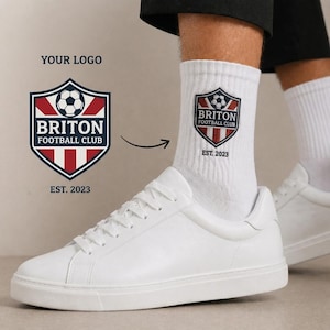 Può includere: Scarpe sportive bianche e calzini bianchi con il logo di un club di calcio. Il logo include un pallone da calcio, il testo "BRITON FOOTBALL CLUB" e l'anno "EST. 2023". Il logo è rosso, bianco e blu.