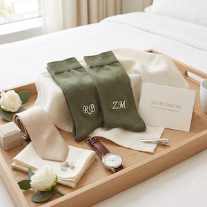 May include: A pair of olive green socks embroidered with initials RB and ZM, a beige necktie, a white pocket square, silver cufflinks, a silver tie bar, a wristwatch with a brown leather strap, and a card reading TO MY GROOM. I can't wait to marry you.