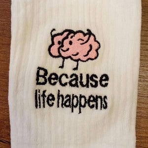 Puede incluir: Un calcet&iacute;n blanco acanalado con un bordado de un cerebro de dibujos animados rosa con cara sonriente y piernas negras delgadas. Debajo del cerebro, el texto "Because life happens" est&aacute; bordado en hilo negro con una fuente negrita y texturizada.