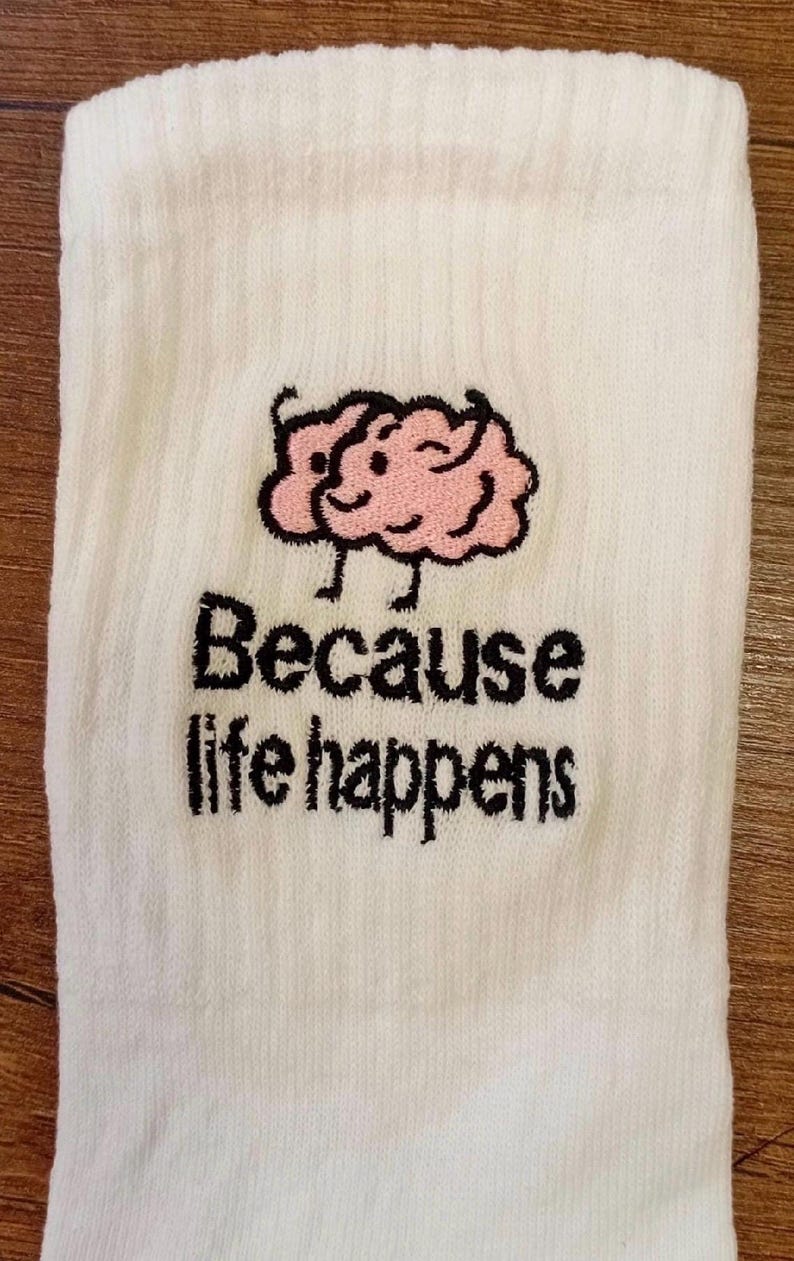 Puede incluir: Un calcet&iacute;n blanco acanalado con un bordado de un cerebro de dibujos animados rosa con cara sonriente y piernas negras delgadas. Debajo del cerebro, el texto "Because life happens" est&aacute; bordado en hilo negro con una fuente negrita y texturizada.