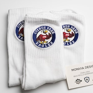 Puede incluir: Tres calcetines blancos con un parche bordado circular que muestra un &aacute;guila marr&oacute;n con una camiseta roja etiquetada CPE y el texto Citrus Park Eagles. Una tarjeta blanca muestra el texto Monvia Design, Your Logo Beautifully Embroidered.