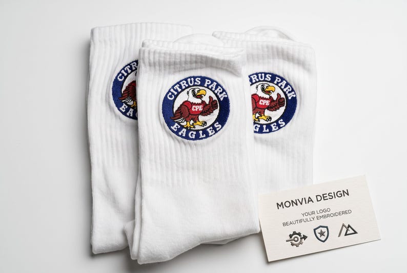 Puede incluir: Tres calcetines blancos con un parche bordado circular que muestra un &aacute;guila marr&oacute;n con una camiseta roja etiquetada CPE y el texto Citrus Park Eagles. Una tarjeta blanca muestra el texto Monvia Design, Your Logo Beautifully Embroidered.