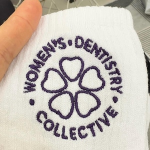 Puede incluir: Un art&iacute;culo de tela blanca con un logotipo circular bordado en color morado. El texto "WOMEN'S DENTISTRY COLLECTIVE" aparece en un c&iacute;rculo alrededor de una forma central similar a una flor, compuesta por cinco p&eacute;talos en forma de coraz&oacute;n.