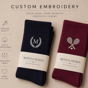 Puede incluir: Dos pares de calcetines acanalados, uno azul marino y otro burdeos, envueltos en una banda de papel con la etiqueta MONVIA DESIGN CUSTOM EMBROIDERY. El calcet&iacute;n azul marino tiene una letra M bordada en blanco dentro de una corona de laurel, mientras que el burdeos muestra dos raquetas de tenis cruzadas y una pelota. El texto superior dice CUSTOM EMBROIDERY, YOUR LOGO, YOUR IDENTITY, PERFECTLY YOURS, con iconos de calidad premium, suavidad, tejido transpirable y durabilidad.
