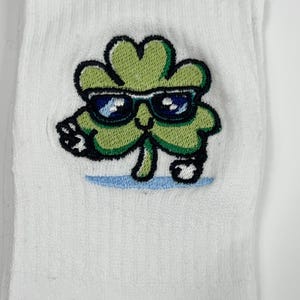 Puede incluir: Un calcet&iacute;n blanco con un bordado de un personaje de tr&eacute;bol de cuatro hojas de color verde claro. El tr&eacute;bol tiene una cara sonriente, usa gafas de sol azul oscuro y est&aacute; posicionado sobre un peque&ntilde;o detalle de l&iacute;nea azul claro.