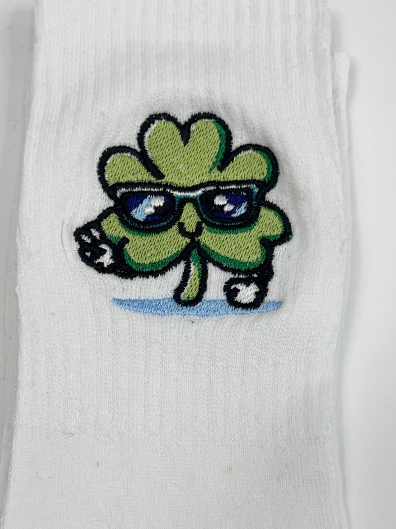 Puede incluir: Un calcet&iacute;n blanco con un bordado de un personaje de tr&eacute;bol de cuatro hojas de color verde claro. El tr&eacute;bol tiene una cara sonriente, usa gafas de sol azul oscuro y est&aacute; posicionado sobre un peque&ntilde;o detalle de l&iacute;nea azul claro.