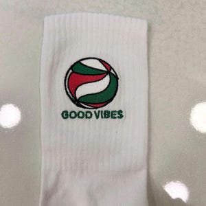 Puede incluir: Un calcet&iacute;n blanco con un gr&aacute;fico bordado de un bal&oacute;n de voleibol en rojo, blanco y verde con un contorno negro. Debajo del bal&oacute;n, el texto GOOD VIBES est&aacute; bordado en letras may&uacute;sculas verdes. El calcet&iacute;n tiene una textura acanalada.