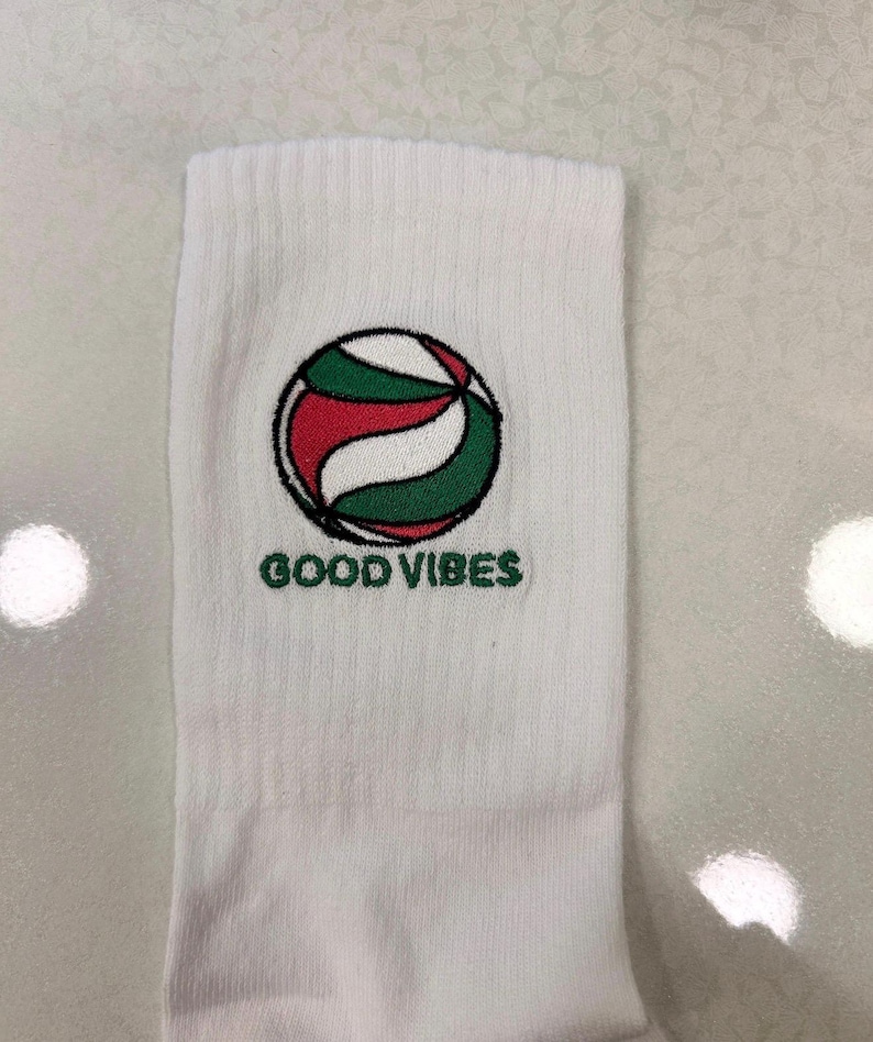 Puede incluir: Un calcet&iacute;n blanco con un gr&aacute;fico bordado de un bal&oacute;n de voleibol en rojo, blanco y verde con un contorno negro. Debajo del bal&oacute;n, el texto GOOD VIBES est&aacute; bordado en letras may&uacute;sculas verdes. El calcet&iacute;n tiene una textura acanalada.