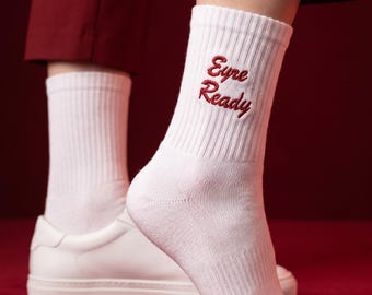 Puede incluir: Un par de calcetines blancos de canalé hasta media pantorrilla con el texto bordado "Eyre Ready" en hilo rojo oscuro. Los calcetines tienen un patrón de punto texturizado. El bordado está situado en el lado exterior de la zona del tobillo.