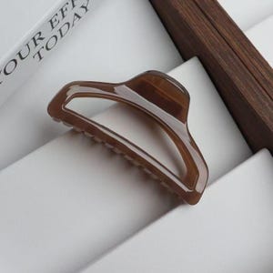 May include: A brown hair claw clip with a curved design. The clip has a matte finish and a row of teeth along the bottom edge. The clip is made of a translucent material and is set against a white background.