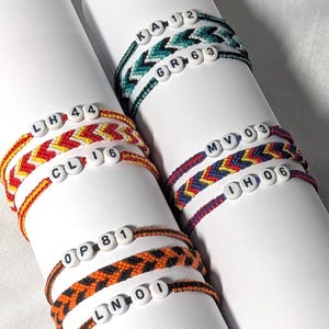 Peut inclure: Une collection de six bracelets d'amitié tissés à motif chevron dans diverses combinaisons de couleurs, notamment bleu sarcelle, rouge, jaune, bleu et orange. Chaque bracelet comporte des perles rondes blanches avec des lettres et des chiffres noirs, tels que KA 12, GR 63, LH 44, CL 16, MV 03, IH 06, OP 81 et LN 01.