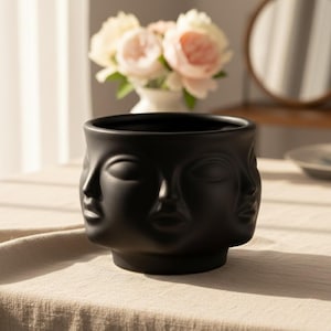 May include: A black ceramic planter with a unique design featuring multiple faces. The faces are sculpted with closed eyes and serene expressions. The planter is round with a wide opening, perfect for displaying plants or flowers. The matte finish adds a touch of elegance.
