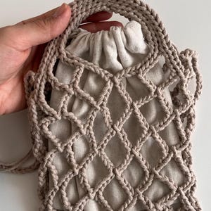 Puede incluir: Un bolso beige tejido a ganchillo con un patrón de diamantes superpuesto a una bolsa con cordón. El bolso tiene un asa y una correa para el hombro. El bolso está hecho de un material de fibra natural.