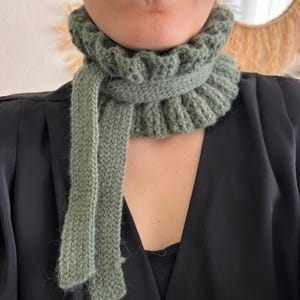 May include: A sage green knitted cowl scarf with a ribbed design and a long, rectangular scarf element. The scarf is made of a soft, textured yarn and is worn around the neck.