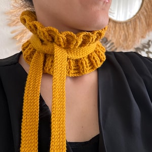 May include: Mustard yellow knitted scarf with a ruffled collar and long ties. The scarf is made of a thick yarn and has a textured appearance. The scarf is a warm and stylish accessory.