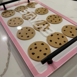 May include: A rectangular serving tray with a white base, a light pink border, and two black metal handles. The surface features a repeating pattern of tan chocolate chip cookie graphics and the white text "STAY" written in a bold, sans-serif font.