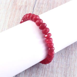 Pode incluir: Uma pulseira de pedras preciosas vermelhas com contas facetadas. A pulseira é exibida em uma superfície cilíndrica branca contra um fundo de grão de madeira clara. As contas são de uma cor vermelha profunda.