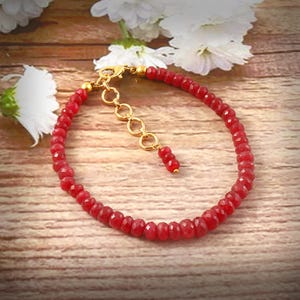Puede incluir: Una pulsera de cuentas rojas con un cierre y una cadena extensora dorados. La pulsera está hecha de pequeñas cuentas rojas facetadas. El fondo es una superficie de madera con flores blancas.